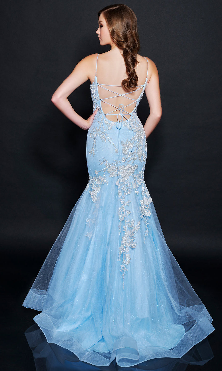 A woman faces away, wearing the Nina Canacci 9120 long formal dress in light blue with floral lace, mermaid silhouette, and lace-up back against a dark background.